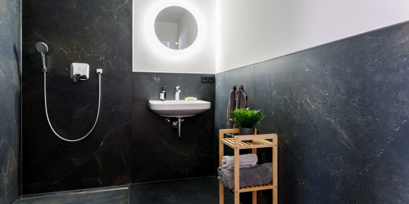 Modernes Bad mit bodengleicher Dusche, dunklen Steinfliesen, beleuchtetem Rundspiegel, Waschtisch und Holzregal mit Handtüchern und Pflanze.