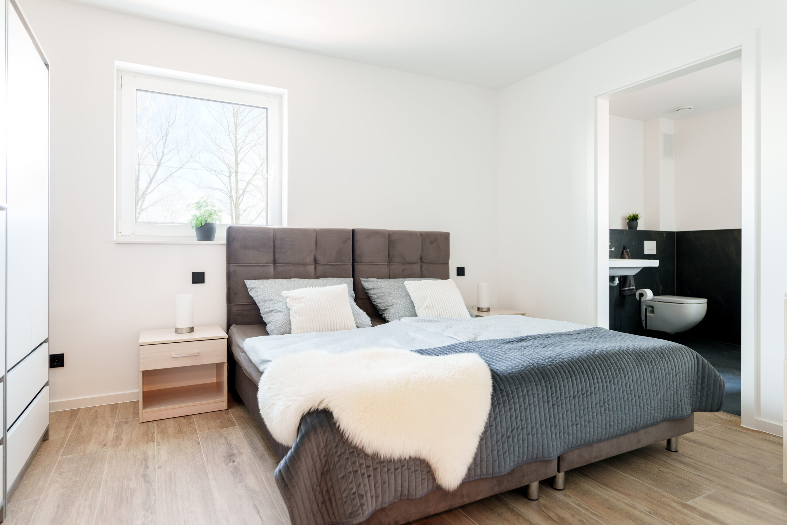 Helles Schlafzimmer mit Doppelbett, Holzoptik-Boden, zwei Nachttischen, Fenster mit Pflanze und direktem Zugang zu einem modernen Badezimmer.